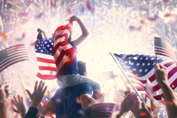 A child sits on their father's shoulders with U.S. flags and red, white, and blue fireworks in the background.