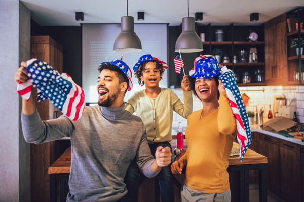 Three people with U.S. flags and red, white, and blue hats stand in a kitchen.