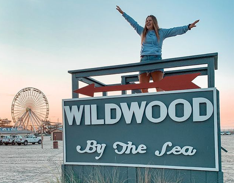 A smiling woman stands on a beach sign with a ferris wheel in the background.