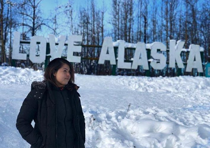 An au pair stands in front of a sign that says "Love Alaska," with snow covering the ground.