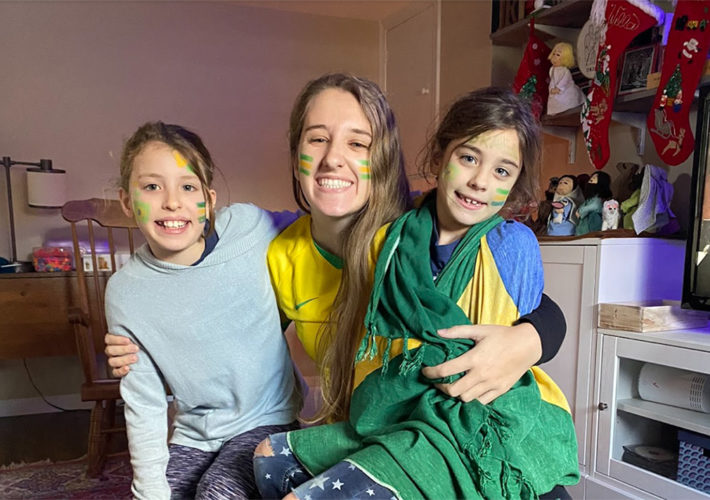 Female au pair poses with two school-aged girls with Brazilian flag-inspired face paing.