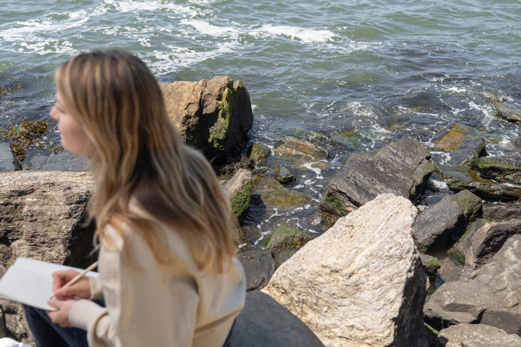 Woman sitting on rocks by the water and journaling.