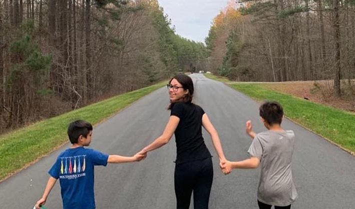 A young woman holds hands with two boys on a road surrounded by trees.