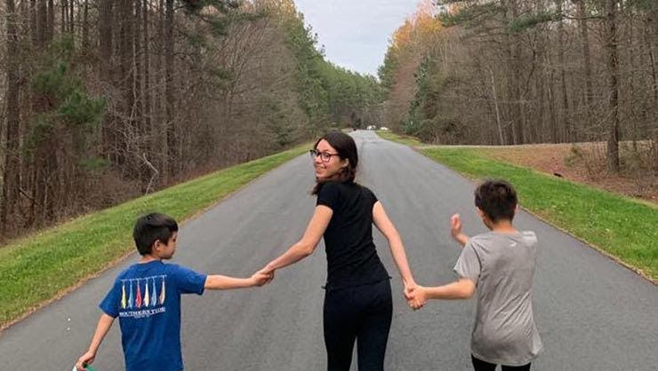 A young woman holds hands with two boys on a road surrounded by trees.