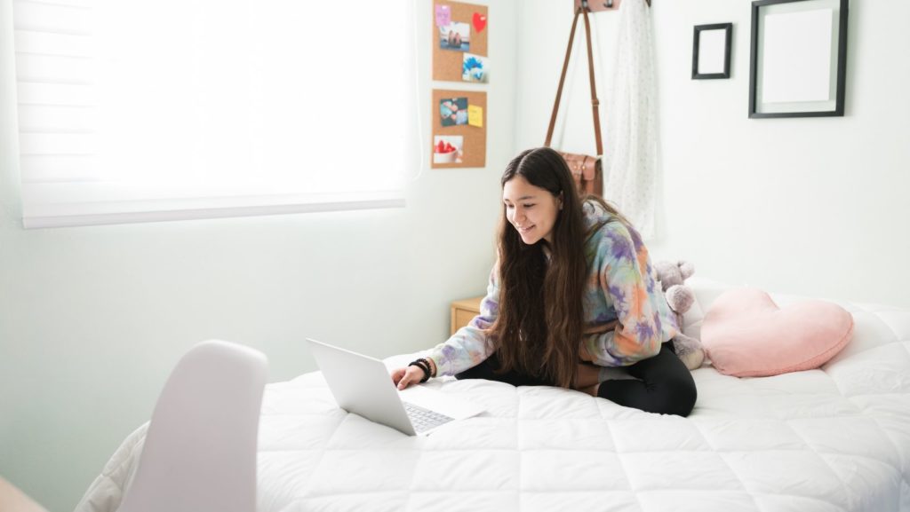 Young female au pair sits on her bed looking at a laptop computer.